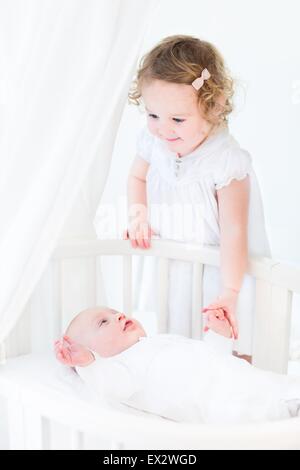 Happy toddler girl with curly hair wearing a pink dress holding her baby brother's hand in a white sunny bedroom Stock Photo