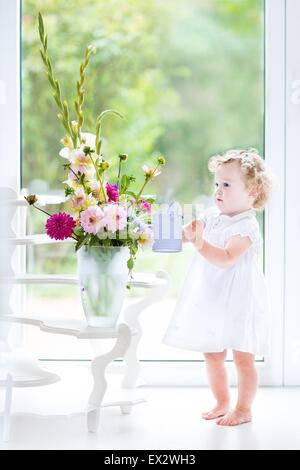 Cute toddler girl in a white dress watering flowers next to a big window with garden view Stock Photo