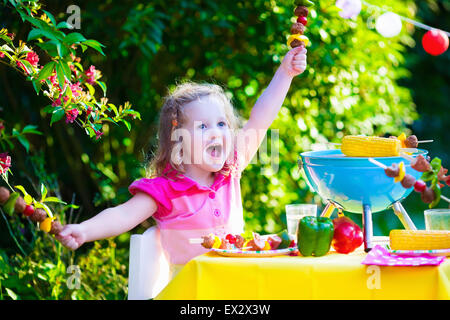 Children grilling meat. Family camping and enjoying BBQ. Little girl at barbecue preparing steaks, kebab and corn. Stock Photo