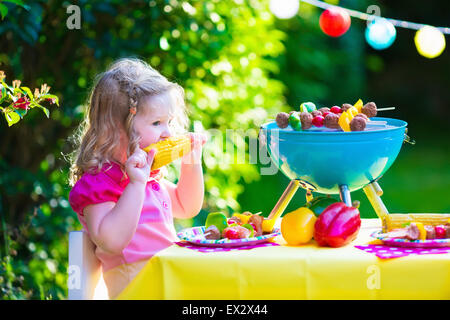 Children grilling meat. Family camping and enjoying BBQ. Little girl at barbecue preparing steaks, kebab and corn. Stock Photo