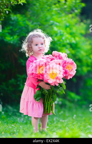 Little cute girl with peony flowers. Child wearing a pink dress playing in a summer garden. Kids gardening. Stock Photo