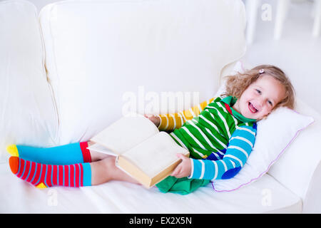Little girl reading a book relaxing on a white couch. Kids read books at home or preschool. Children learning and doing homework Stock Photo