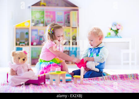 Kids playing with doll house and stuffed animal toys. Children sit on a pink rug in a play room at home or kindergarten. Stock Photo