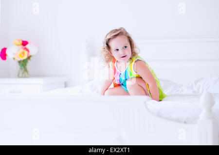 Children jump on a bed. Cute little girl jumping and dancing in a sunny white bedroom. Kids room with garden view window. Stock Photo