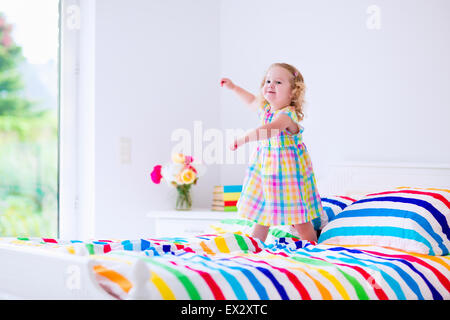 Children jump on a bed. Cute little girl jumping and dancing in a sunny white bedroom. Kids room with garden view window. Stock Photo