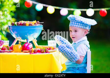 Children grilling meat. Family camping and enjoying BBQ. Little boy at barbecue preparing steaks, kebab and corn. Stock Photo