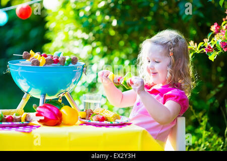 Children grilling meat. Family camping and enjoying BBQ. Little girl at barbecue preparing steaks, kebab and corn. Stock Photo