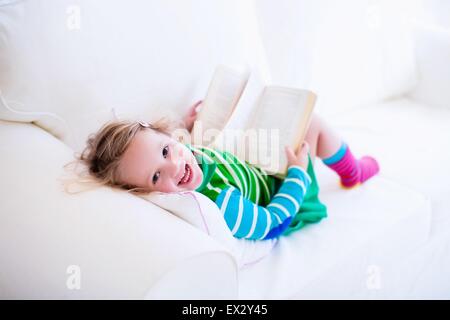 Little girl reading a book relaxing on a white couch. Kids read books at home or preschool. Children learning and doing homework Stock Photo
