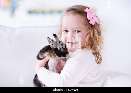 Child playing with a real rabbit. Kids play with pets. Little girl holding bunny. Children and animals at home or preschool. Stock Photo