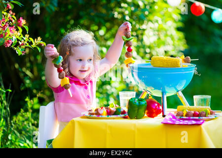 Children grilling meat. Family camping and enjoying BBQ. Little girl at barbecue preparing steaks, kebab and corn. Stock Photo