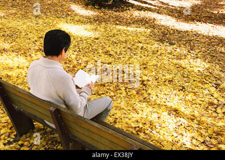 Senior Japanese man sitting on a bench with a book in a city park in Autumn
