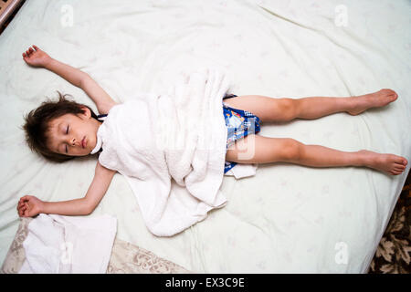 Mixed race, Japanese-English child, boy, 3-4 year old, sleeping on bed with towel over him. Arms outstretched on bed over head, eyes closed. Stock Photo