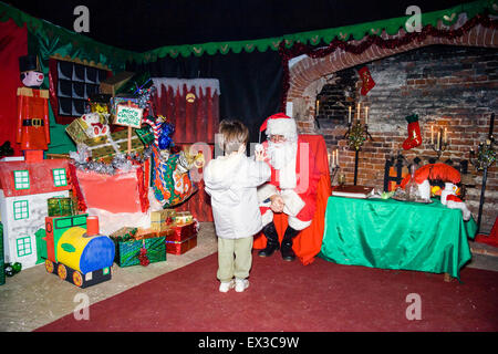 Boy in Santa's Grotto being questioned by Father Christmas. Caucasian child, 3-4 years old, wears winter coat. Santa surrounded by presents. Stock Photo