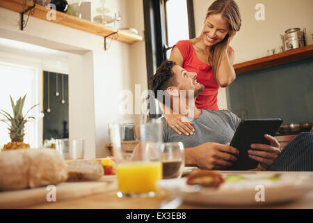 Happy young couple in their kitchen in morning. Man sitting at breakfast table with a digital table with woman standing next to Stock Photo