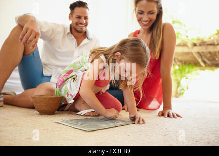 Shot of little girl drawing picture with her parents sitting behind her in living room. Family sitting on floor with daughter pa Stock Photo