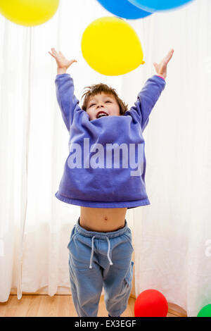 Caucasian child, boy, playing indoors with colored balloons. 6-7 year old enjoying throwing balloons upwards against background of white curtains.