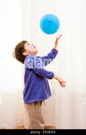 Caucasian child, boy, playing indoors with colored balloons. 6-7 year old enjoying hitting floating balloons against a background of white curtains.