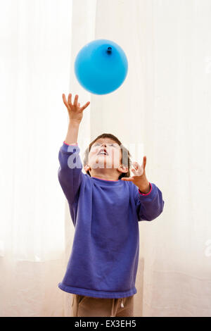 Caucasian child, boy, playing indoors with colored balloons. 6-7 year old enjoying throwing balloons upwards against background of white curtains.