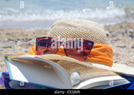 Straw hat sunglasses and a book on the beach with sea in backgound on a sunny day Stock Photo