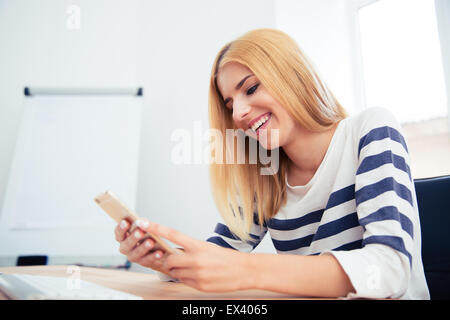 Happy young woman sitting at the table and using smartphone in office Stock Photo