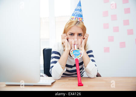 Young girl sitting at the table with party hat in office and blows whistle Stock Photo