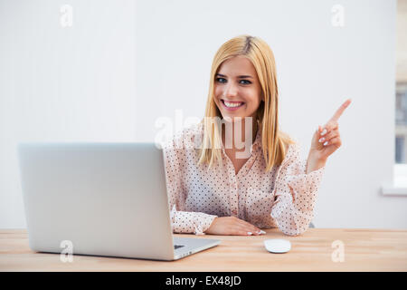 Smiling businesswoman sitting at the table with laptop and poing finger away over gray background. Looking at camera Stock Photo
