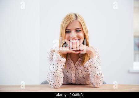 Smiling blonde businesswoman sitting at the desk over gray background and looking at camera Stock Photo