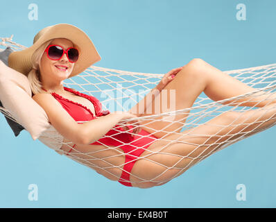 Beautiful woman in a red swimsuit wearing a stylish hat and lying in a hammock on blue background Stock Photo