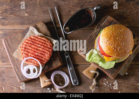 Fresh homemade burger on little cutting board and raw cutlet and sliced onion, served with ketchup sauce and meat fork over wood Stock Photo