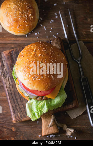 Fresh homemade burger on little cutting board with meat fork over wooden table. Dark rustic style. Top view. Selective focus Stock Photo