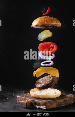 Flying ingredients for homemade burger on little wooden cutting board over dark background. Stock Photo