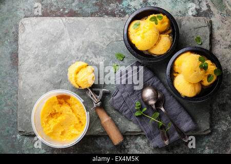 Mango ice cream sorbet with mint leaves in black bowls, Spoon for ice cream and plastic container on stone slate background Stock Photo