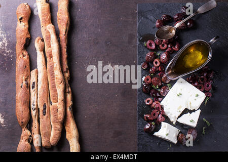 Whole and sliced black olives and block of feta cheese with olive oil and homemade grissini bread sticks on black slate over dar Stock Photo