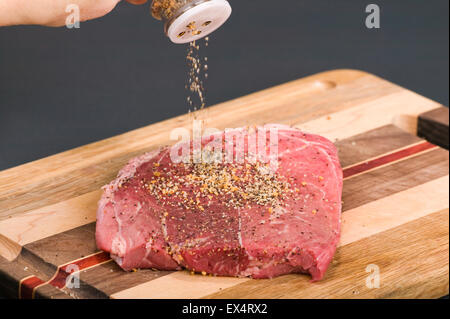 Meat tenderizer from a bottle being sprinkled on a steak sitting on a wooden cutting board, in a studio setting Stock Photo