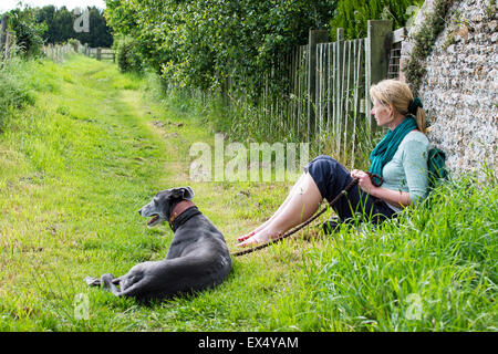 Resting during a dog walk.  Woman and greyhound sitting against a wall near a country footpath. Stock Photo