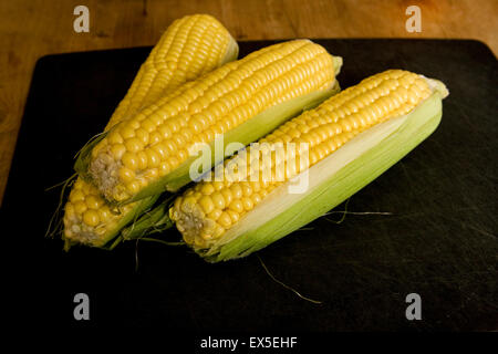 Three fresh corn on the cob arranged on a black chopping board  shot from above. A rustic and natural food presentation with a dark background. Stock Photo