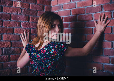 Scared young woman standing in corner of brick wall and looking at camera Stock Photo