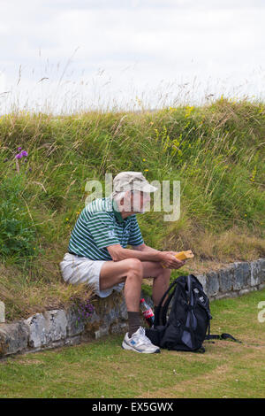 Senior man sitting on wall holding paperback book at the Iron Age hill fort hillfort at Old Sarum, near Salisbury, Wiltshire UK in July Stock Photo