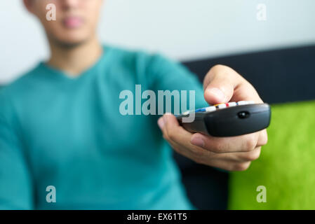 Asian young adult man watching TV and changing channel with remote control. Narrow focus on buttons and hand in foreground, crop Stock Photo