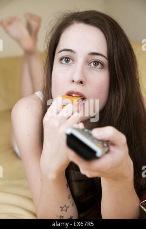 Young woman watching TV on the couch with chips and the remote control in hand Stock Photo