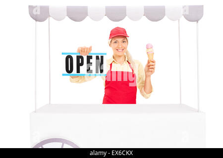 Young female vendor holding an ice cream cone and an open sign behind an ice cream stand isolated on white background Stock Photo