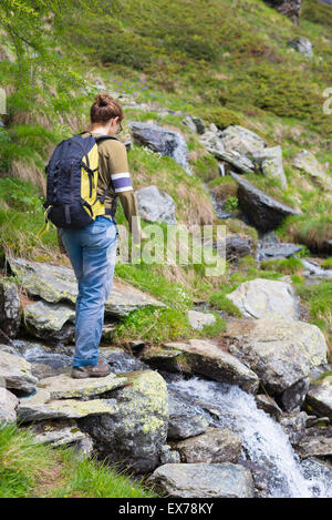 Female hiker with backpack crossing little stream in the mountains. Summer adventures in the italian Alps. Stock Photo