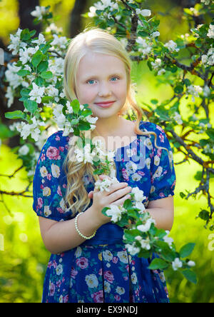 Adorable toddler girl with curly hair and flower crown wearing a magic fairy costume with a blue dress and angel wings playing in a sunny blooming fruit garden with cherry blossom and apple trees. Stock Photo