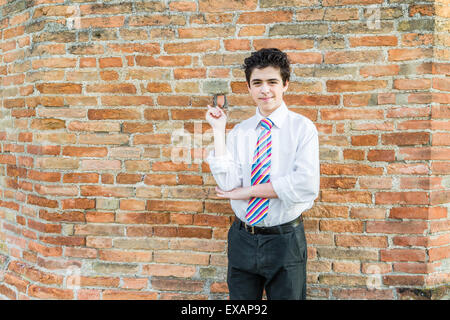 Handsome Caucasian boy wearing a white shirt and a regimental tie with red, fuchsia, orange, blue, indigo and white stripes is pointing something in front of a brick wall at sunset Stock Photo