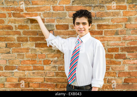 Handsome Caucasian boy wearing a white shirt and a regimental tie with red, fuchsia, orange, blue, indigo and white stripes is showing in front of a brick wall at sunset Stock Photo