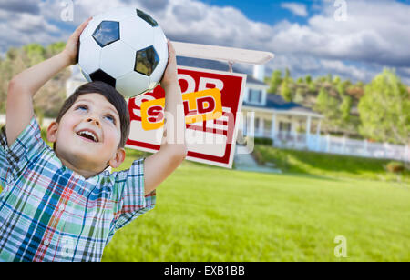 Playful Young Boy Holding Soccer Ball In Front of House and Sold For Sale Real Estate Sign. Stock Photo