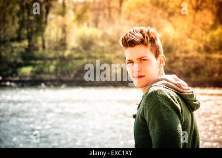 Head and shoulders length of contemplative light brown haired teenage boy wearing green hooded-shirt beside picturesque river or Stock Photo