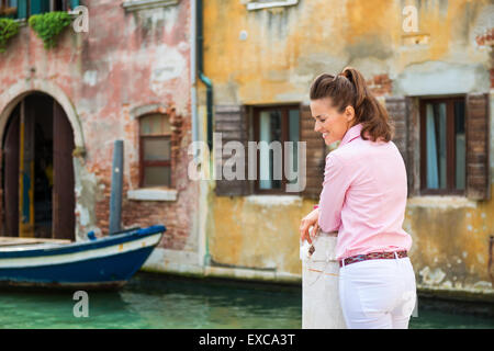 An elegant tourist is standing and relaxing near one of Venice's many canals, looking down while smiling. Stock Photo