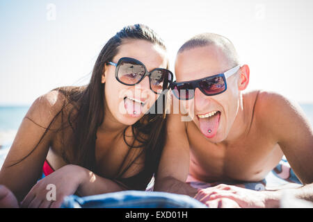couple lying on beach toowl in summertime using smartphone showing tongue piercing Stock Photo