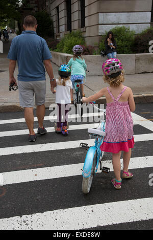 Young children on bicycles crossing the street with an adult  A rear view  New York USA Stock Photo